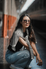 Confident young woman with long, dark hair and sunglasses eagerly awaits her journey in a bustling train station, exuding independence and style