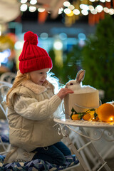 A little girl, wearing a bright red hat, beams with  a fluffy white rabbit, surrounded by the...