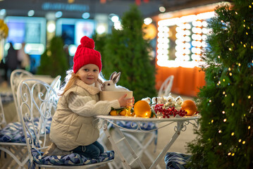 A little girl, wearing a bright red hat, beams with  a fluffy white rabbit, surrounded by the...