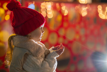A little girl, wearing red hat,  blows playfully on her palm, sending sparkling snowflakes scattering through the air.
