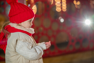 A little girl, wearing red hat,  blows playfully on her palm, sending sparkling snowflakes scattering through the air.