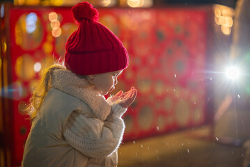 A little girl, wearing red hat,  blows playfully on her palm, sending sparkling snowflakes scattering through the air.