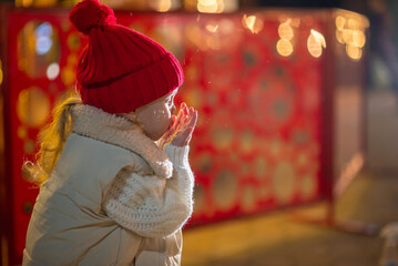 A little girl, wearing red hat,  blows playfully on her palm, sending sparkling snowflakes scattering through the air.