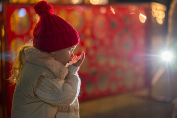 A little girl, wearing red hat,  blows playfully on her palm, sending sparkling snowflakes scattering through the air.