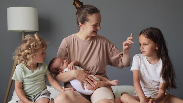 Adorable young woman with bun hairstyle sitting on bed with her little children kids meeting small tiny sibling at home sisters looking at child while posing with mommy