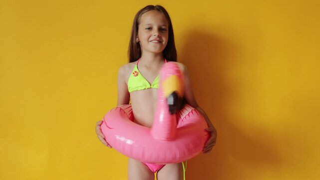 Overjoyed little girl in flamingo rubber ring around waist isolated over yellow background dancing with raised arms playing on summer tropical resort enjoying summer vacation