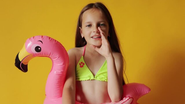 Overjoyed little girl in flamingo rubber ring around waist isolated over yellow background keeps hand near mouth speaking loud making announcement about tropical resort