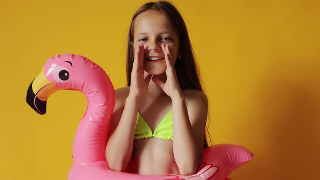 Cheerful adorable little girl in flamingo rubber ring around waist screaming loud with hands near mouth announcing making advertisement isolated over yellow background