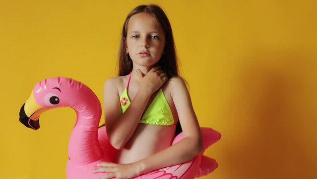 Sick ill little girl in flamingo rubber ring around waist isolated over yellow background swimming for long time in the sea or pool coughing having flu symptoms