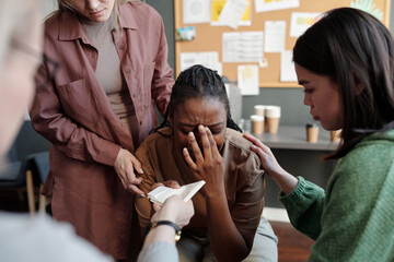 Two young women comforting crying African American girl and keeping hands on her shoulders while...