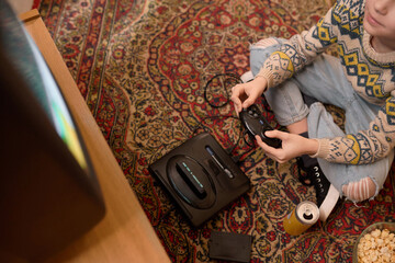 Cropped top view of Caucasian teenage boy in patterned sweater and jeans holding a retro video game controller while sitting on a vintage carpeted floor, next to a drink and popcorn bowl © pressmaster