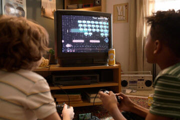 Back view of two kids absorbed in playing a retro video game on CRT TV, sitting on a cozy living...