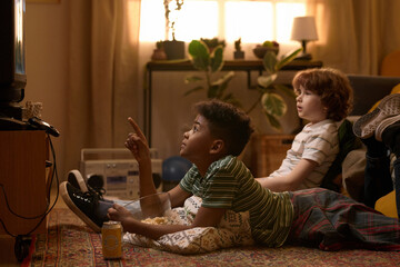 Two multiethnic schoolkids lying on floor, one pointing at TV while watching exciting movie in cozy retro-styled living room with relaxed atmosphere and snacks like popcorn and soda