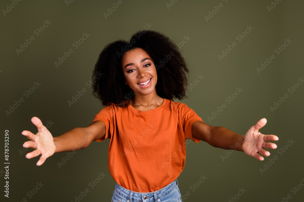 Poster Photo of kind positive woman with perming coiffure dressed orange shirt stretching hands to hug you isolated on dark khaki background