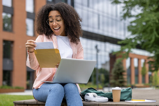 Happy student receives letter of approval sitting on university campus bench with laptop. Joyful reaction to good exam results or enrollment confirmation while studying outdoors.