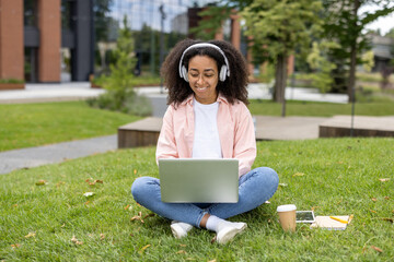 Young woman wearing headphones using laptop in outdoor park setting. She sits cross-legged on grass with coffee cup and notebook nearby, enjoying peaceful environment and technology.
