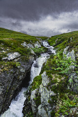 A rocky mountainside in Norway features a stream of water flowing down, creating a cascading waterfall under a gray, cloudy sky