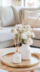 A vase of white roses sits on a gold tray in front of two white candles and a diffuser on a glass coffee table. The table is in front of a white couch with a patterned pillow