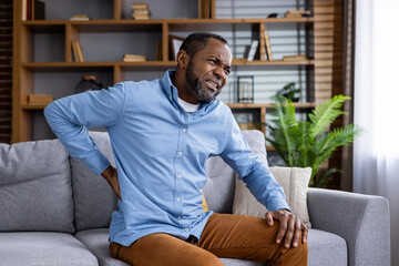 Middle-aged man experiencing back pain while sitting on comfortable couch in modern living room. Man grimacing in discomfort, holding back, in need of relief. Cozy, well-lit home interior setting.