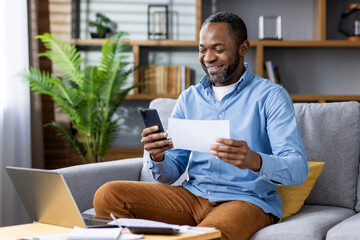 Smiling man working remotely from home, using smartphone and reviewing document while sitting on sofa with laptop and notepad. Concept of remote work home office, happiness, productivity, technology.
