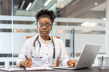 Confident female doctor working at desk in modern clinic. Doctor is using laptop and writing notes, demonstrating professionalism and dedication to medical practice.