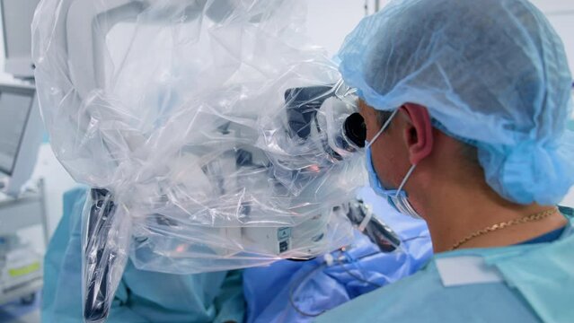 Rear view of a male doctor wearing mask and cap. Medic looks at microscope performing operation. Close up.