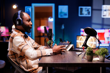Side view portrait of african american male talk show host speaking with his online audience during a live podcast recording from his home studio, he wears wireless headphones.