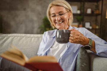 Mature woman sit in the living room read a book and drink coffee