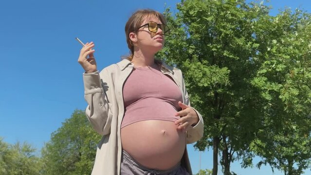 Young pregnant woman is smoking cigarette in summer park