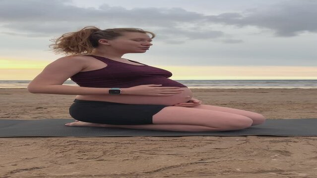 Young pregnant active woman doing exersice outdoors, stratching her body and meditate on the sea, beach at sunset in the evening
