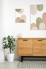 Interior of stylish room with wooden dresser, houseplant and pictures on white wall