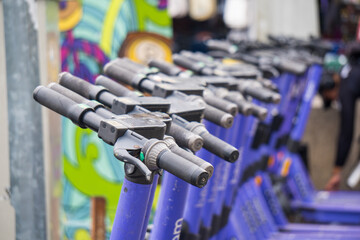 A row of purple and black electric scooters with black handlebars are parked in front of a colorful mural.