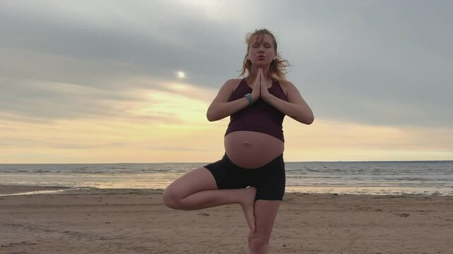 Young pregnant active woman doing exersice outdoors, stratching her body and meditate on the sea, beach at sunset in the evening