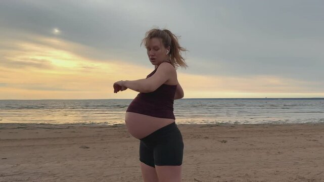 Young pregnant active woman doing exersice outdoors, stratching her body and meditate on the sea, beach at sunset in the evening