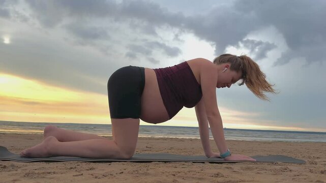 Young pregnant active woman doing exersice outdoors, stratching her body and meditate on the sea, beach at sunset in the evening