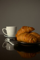 Freshly baked croissants are served on a dark plate alongside a white coffee cup and saucer on a reflective table to create a cozy and inviting breakfast scene that captures the simplicity of morning.