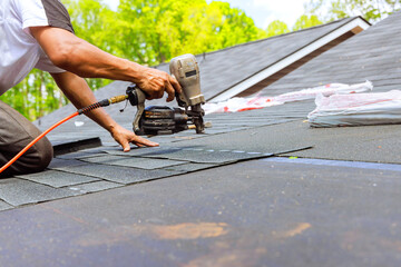 Construction roofer operating an air pneumatic nail gun installed new bitumen shingles on roof