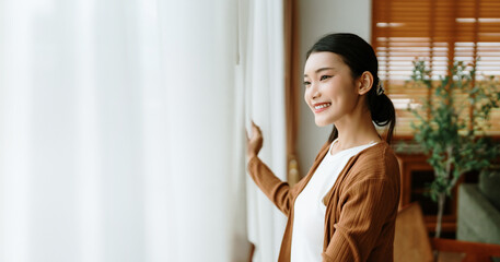 Happy Asian woman standing by the window and looking outside the living room enjoying the bright sunshine