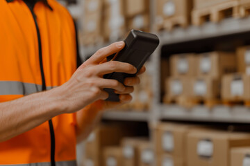 Warehouse worker in orange vest using handheld device for inventory management in a storage facility, surrounded by shelves of boxes.