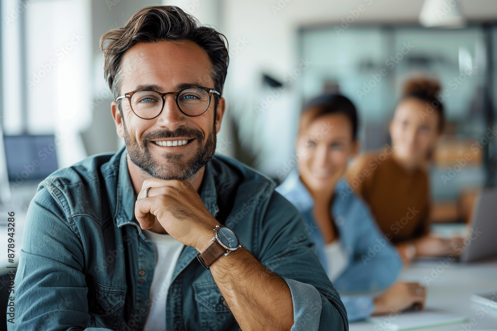 Poster smiling man with glasses sitting at a table in office meeting with colleagues working in background