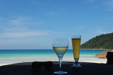 Two glasses of drinks, one is beer and the other is white wine against a scenic white sandy beach, a mountain and blue sky background.