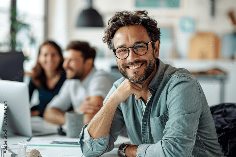 Poster smiling man with glasses sitting in office meeting with team working behind him at table