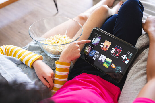 Diverse female friends on couch watching movie on tablet and eating popcorn at home