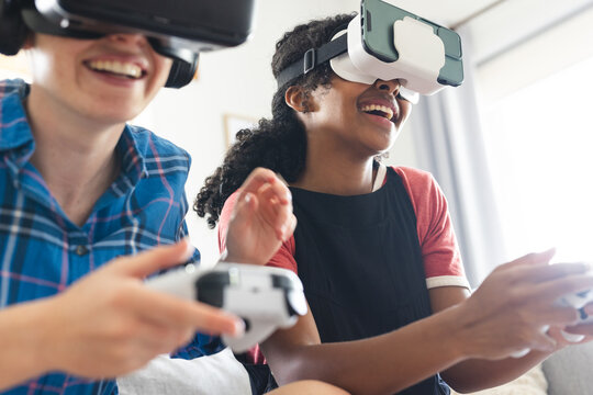 Happy diverse female friends wearing vr headsets and playing video games at home