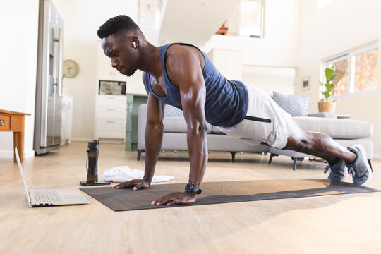 Fit african american man exercising at home, doing push ups and using laptop