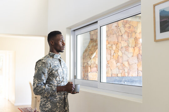 Thoughtful african american male soldier at home holding coffee, looking out of window, copy space