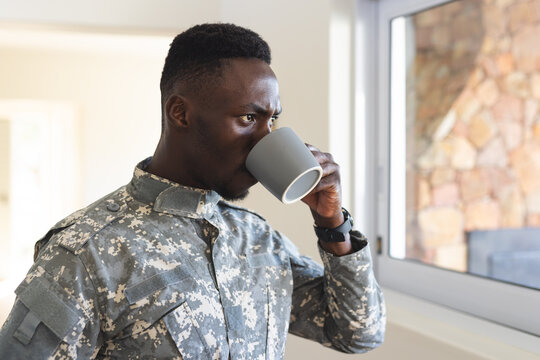 Thoughtful african american male soldier at home holding coffee and looking out of window
