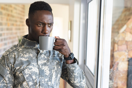 Thoughtful african american male soldier at home holding coffee and looking out of window