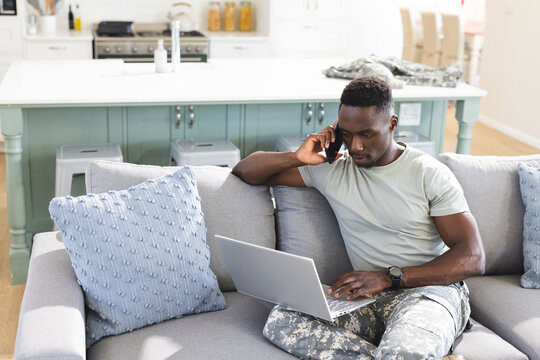 African american male soldier on couch at home using laptop and talking on smartphone, copy space