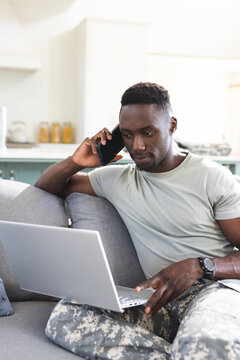 Focused african american male soldier at home using laptop and talking on smartphone, copy space